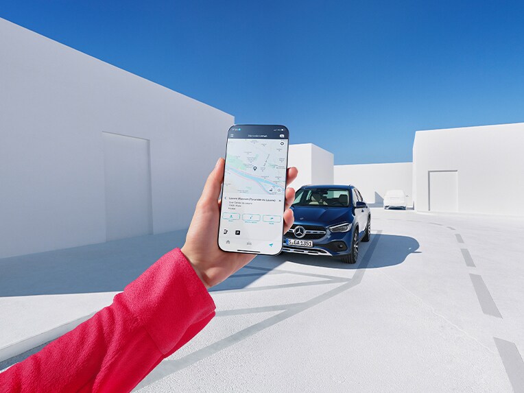 Alguien con un teléfono móvil en el que se puede ver la Mercedes-Benz Service App. En el fondo hay un vehículo Mercedes-Benz detenido.