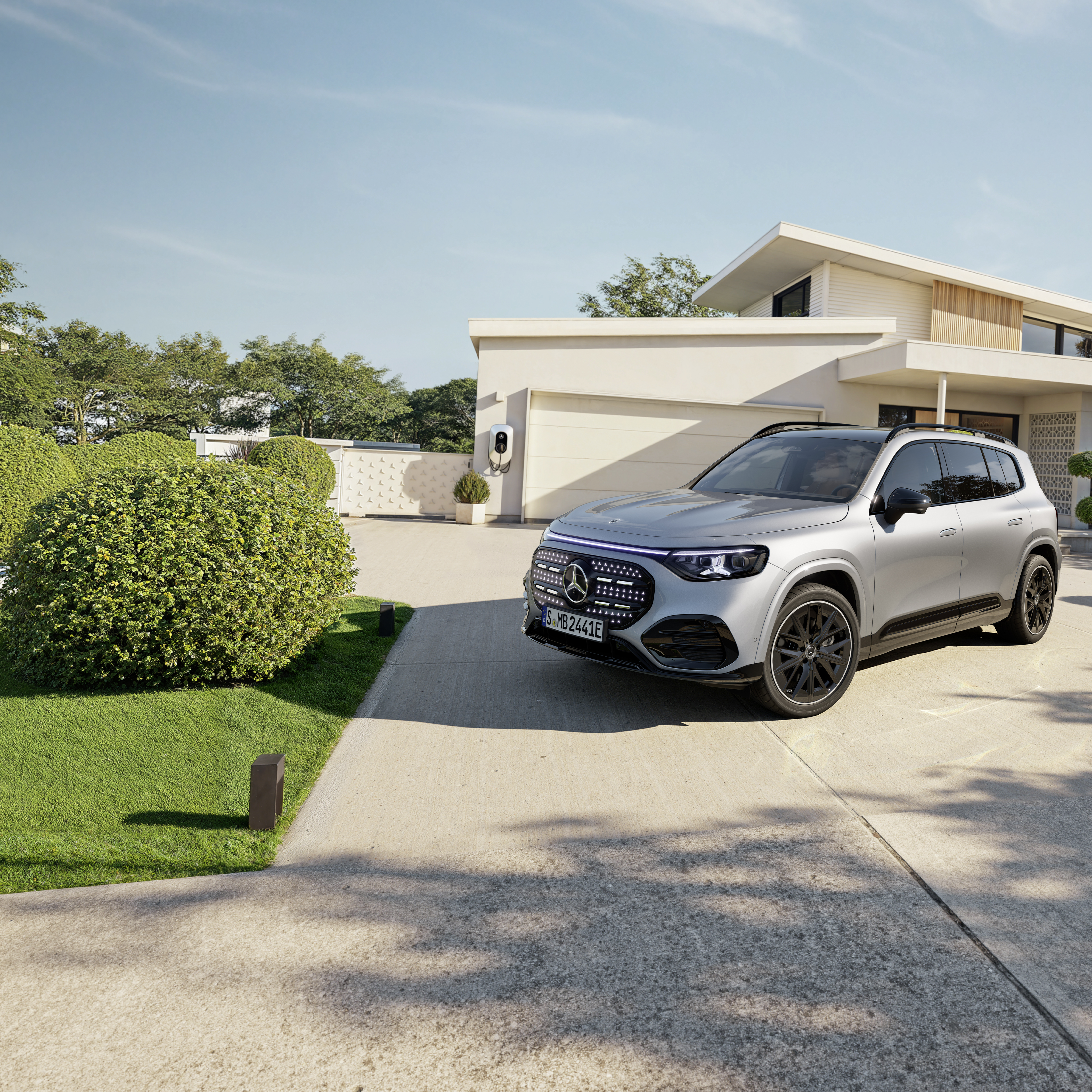 GLB eléctrico | Mercedes-Benz Visual lateral frontal de un GLB gris de Mercedes-Benz aparcado fuera de una casa con un cargador eléctrico instalado