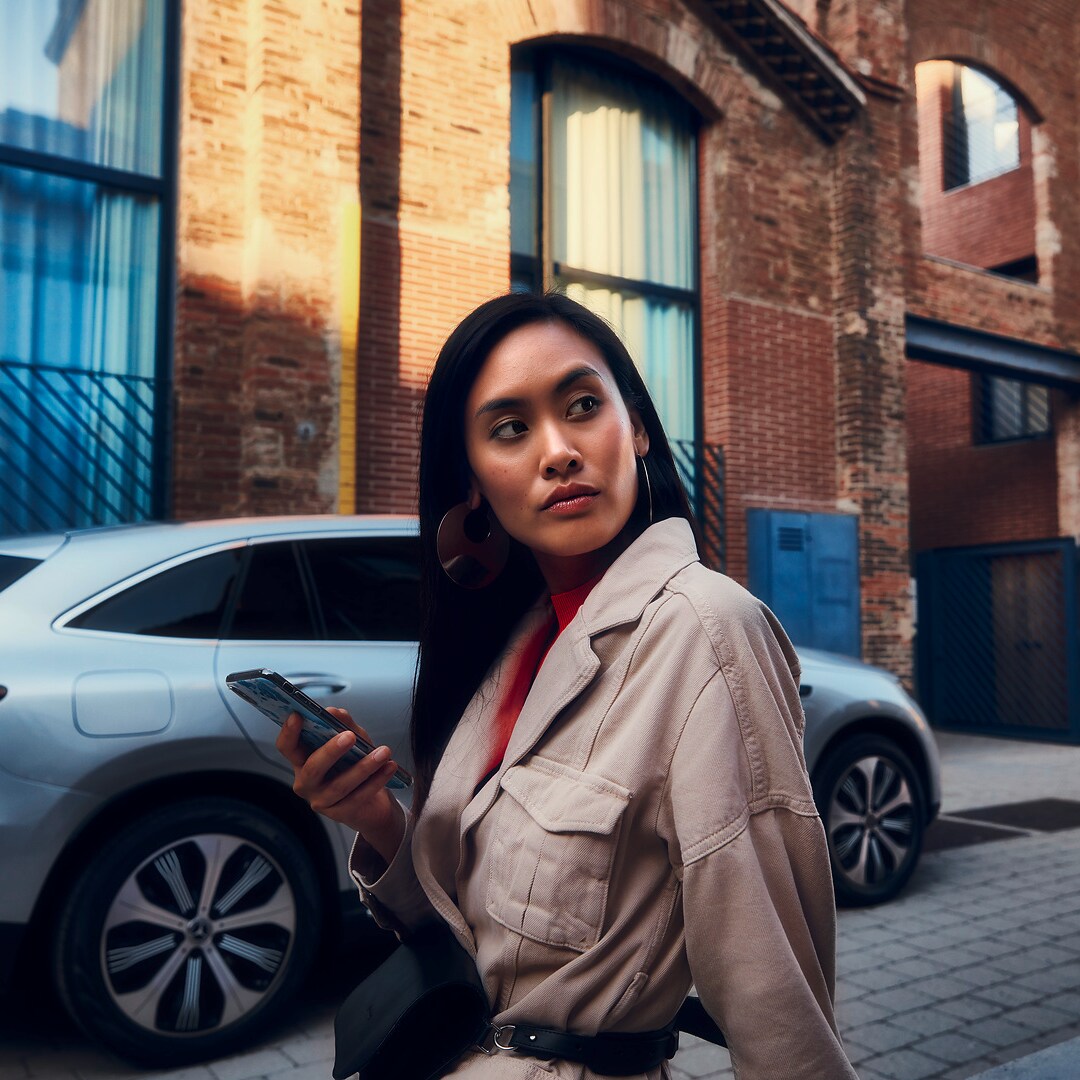 Una mujer se encuentra delante de su Mercedes y sostiene el smartphone en la mano derecha.