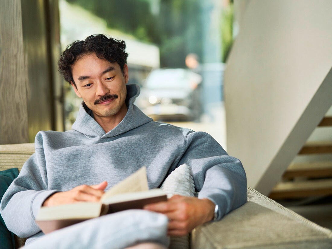 Cliente sentado en casa leyendo un libro. Detrás de él, a través de una ventana, se ve un coche que está siendo revisado por un especialista.