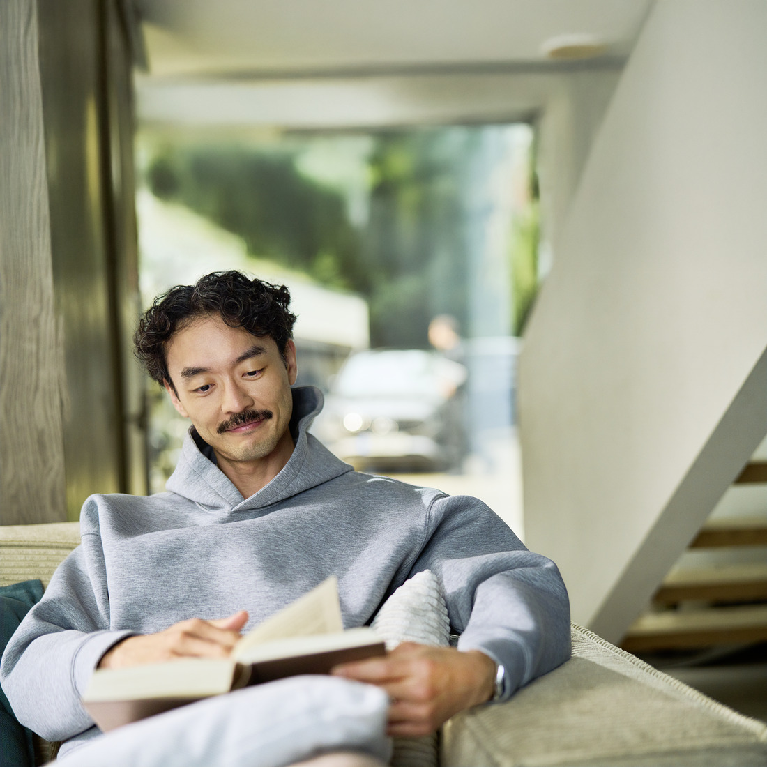 Cliente relajado recibiendo el Servicio Móvil en su domicilio Cliente sentado en casa leyendo un libro. Detrás de él, a través de una ventana, se ve un coche que está siendo revisado por un especialista.