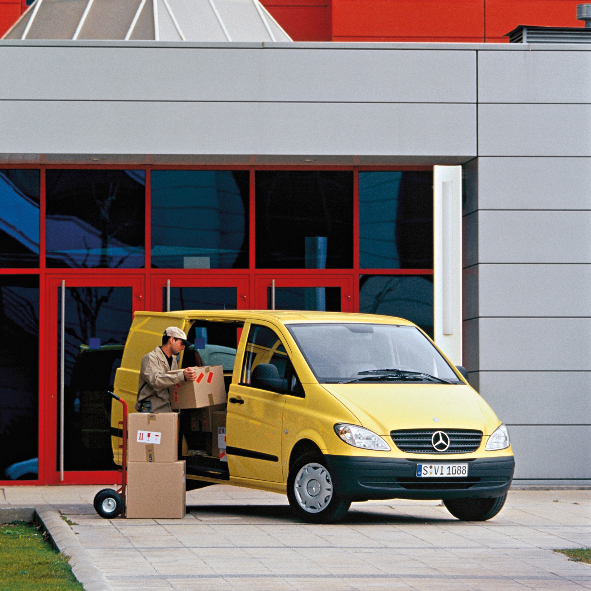 La segunda generación del Mercedes-Benz Vito | Historia de los vehículos industriales | Mercedes-Benz Una Mercedes-Benz Vito amarillo aparcado frente a un edificio, con un trabajador descargando paquetes