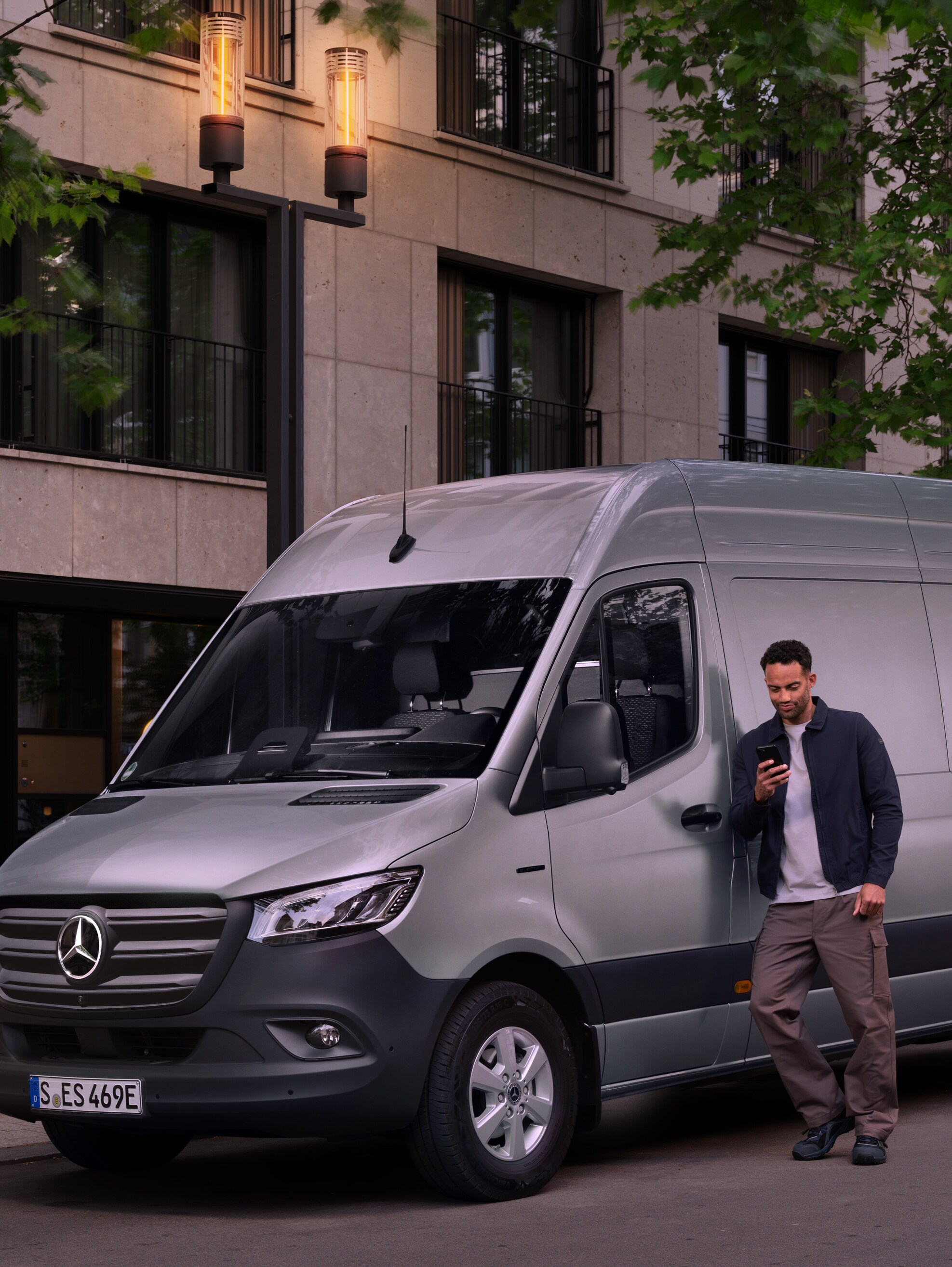 Hombre apoyado en un furgón Mercedes-Benz Sprinter plateado, utilizando su smartphone: asistencia rápida del Servicio de Atención al Cliente de Mercedes Benz.