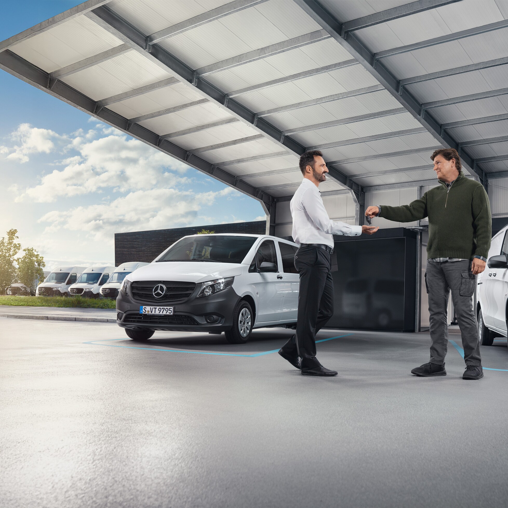 Dos hombres conversando delante de una serie de furgones Mercedes-Benz blancos en una sala.