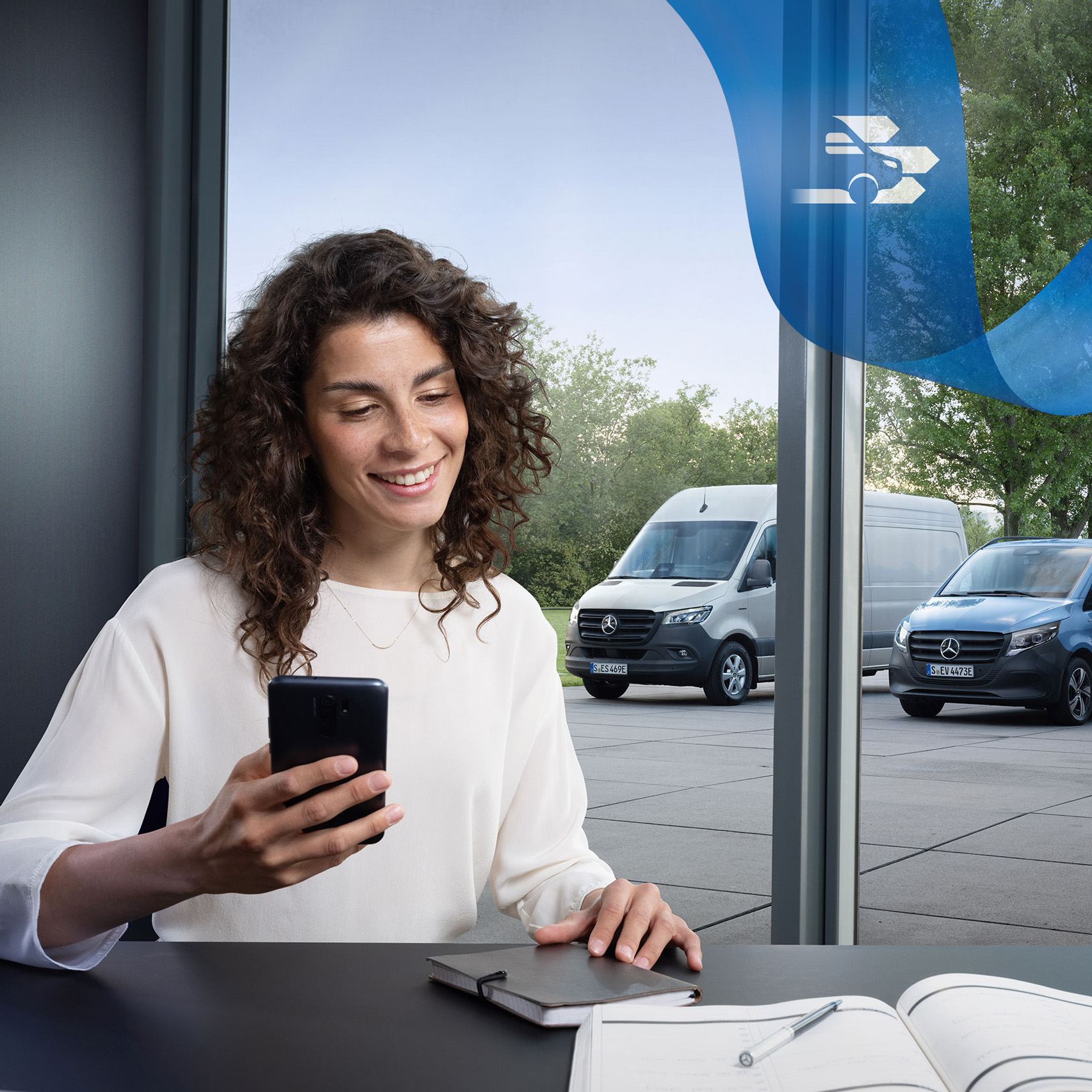 Mercedes-Benz | Cuidado de furgones | Customer Assistance Center Mujer sentada en un escritorio con smartphone y un grupo de furgones Mercedes-Benz de fondo.
