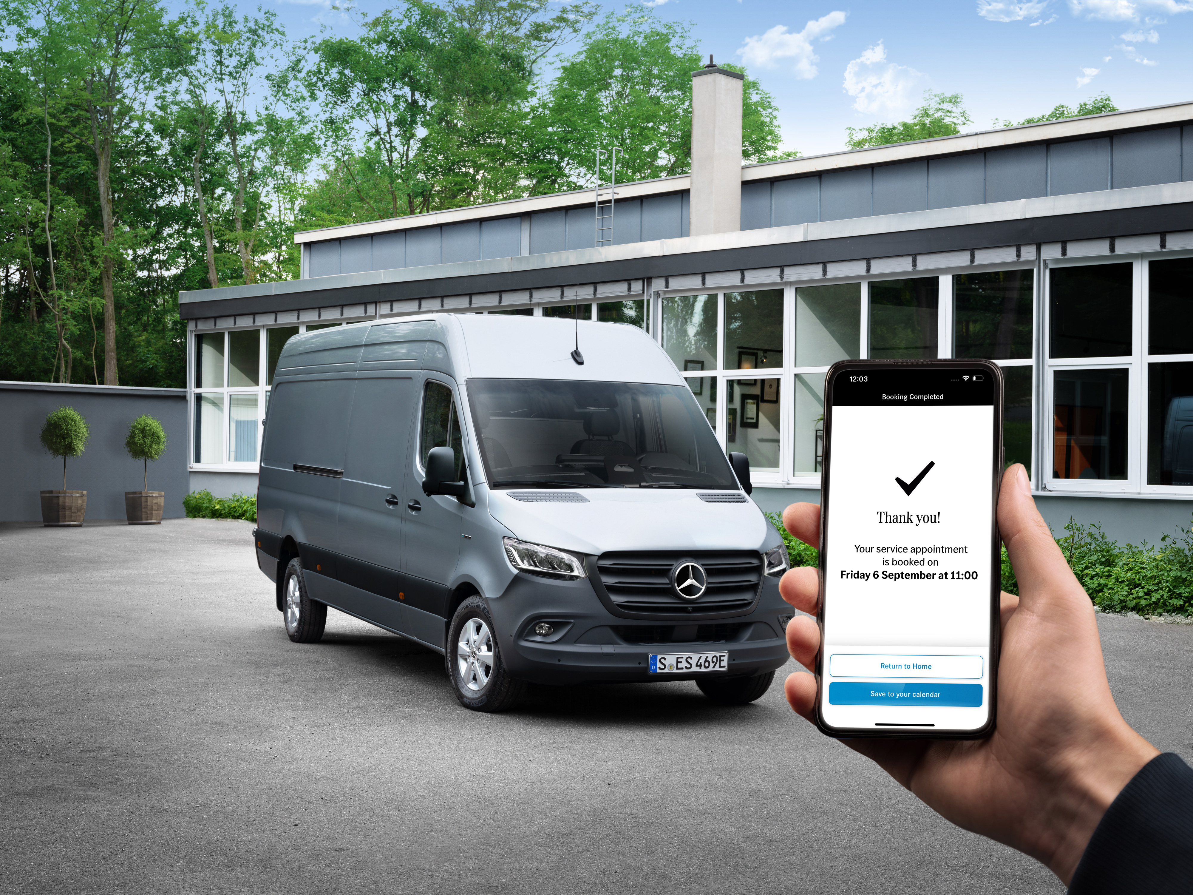 Mercedes-Benz | Conveniencia | Concesionario Oficial Un furgón Mercedes-Benz Sprinter gris delante de una oficina con una persona sosteniendo un smartphone en primer plano.