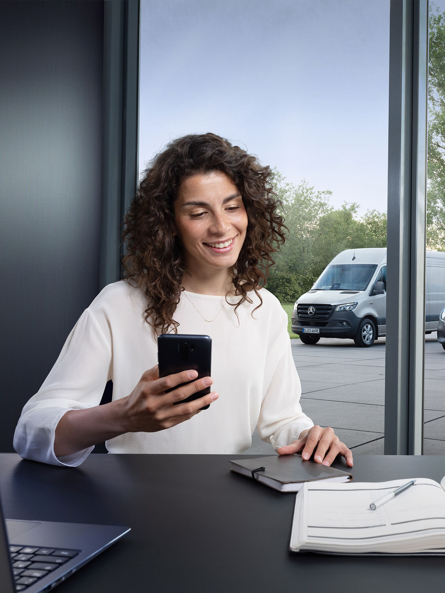Mujer reservando un servicio a través de smartphone con una serie de furgones Mercedes-Benz de fondo.