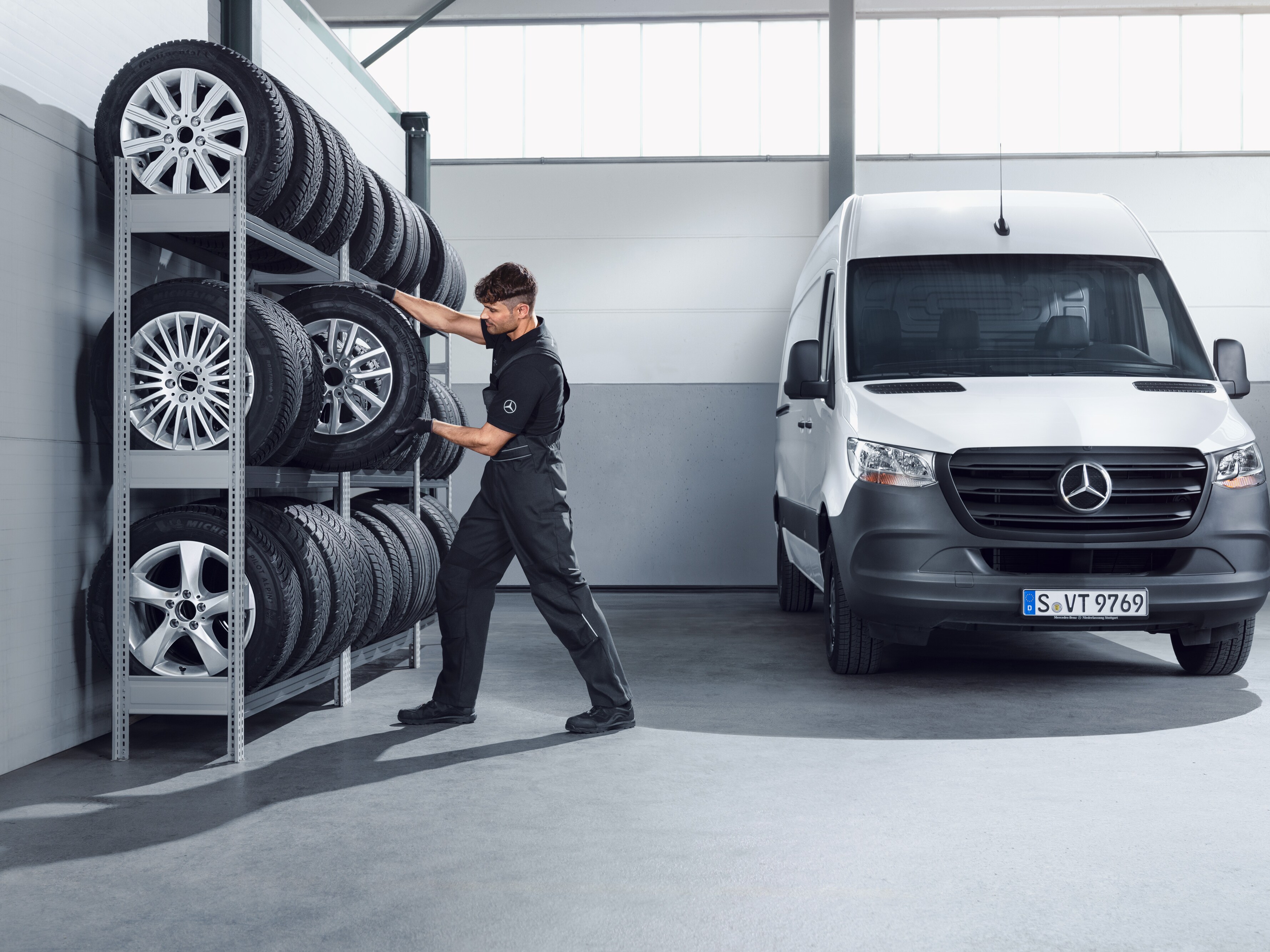 Mecánico eligiendo, de la estantería del taller, ruedas Mercedes-Benz de invierno completas para un Sprinter blanco.