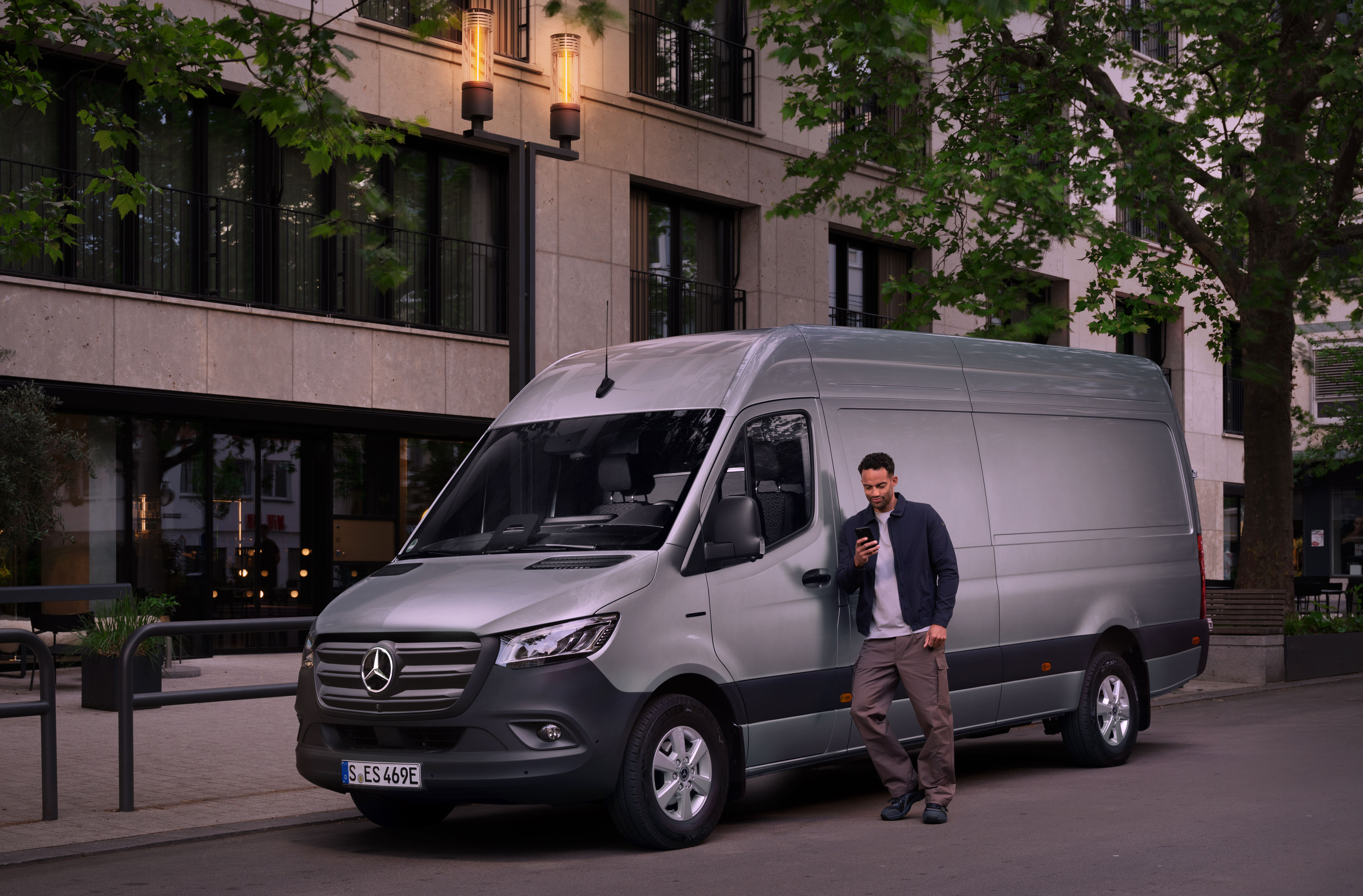 Mercedes-Benz | Reparación | Informe digital de daños Hombre completando un informe de daños en su teléfono móvil, apoyado en un Mercedes-Benz Sprinter plateado.