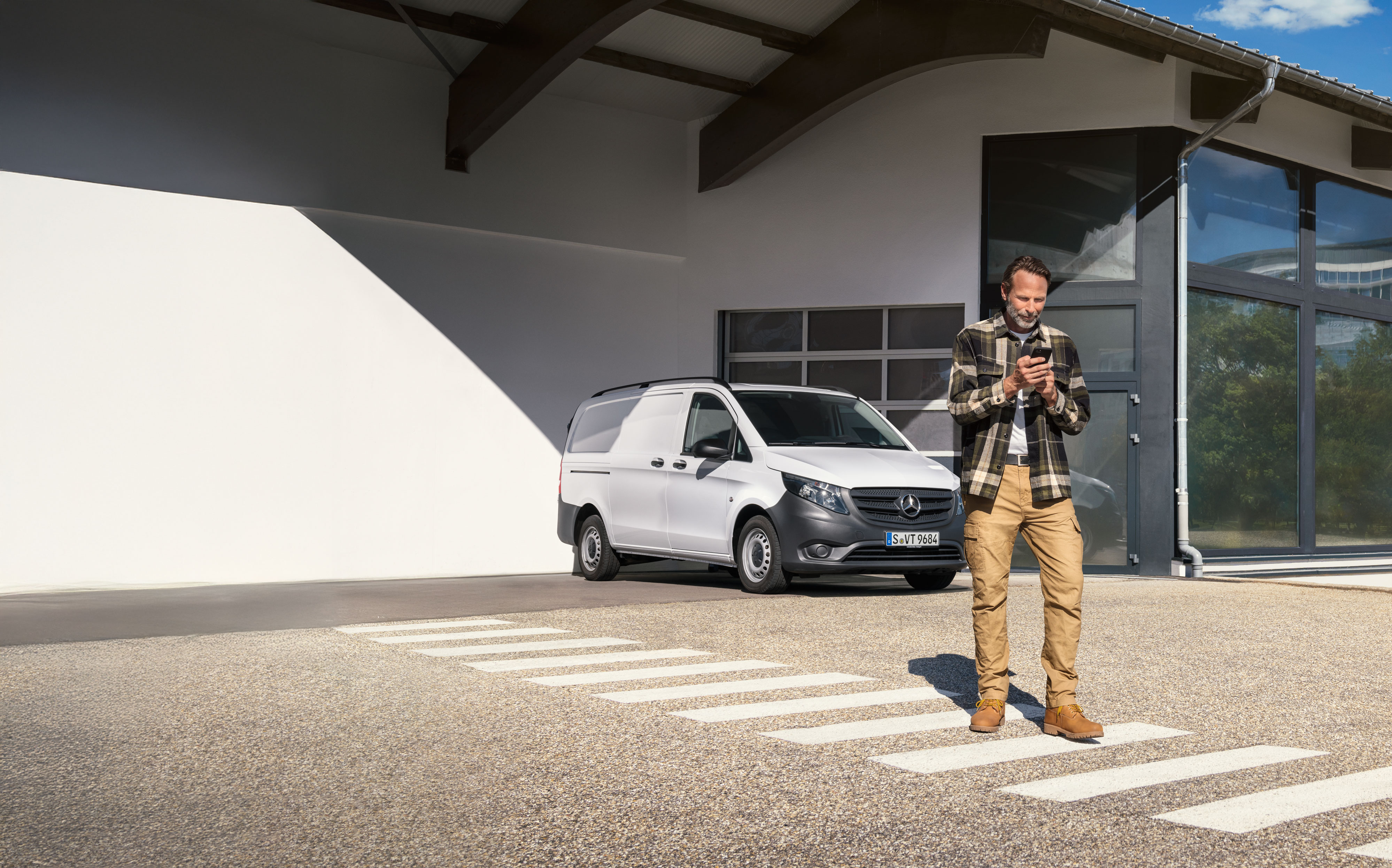 Mercedes-Benz | Mantenimiento | Reserva de cita Un cliente, parado delante de un furgón Mercedes-Benz Vito de color blanco, reservando una cita usando su dispositivo móvil.