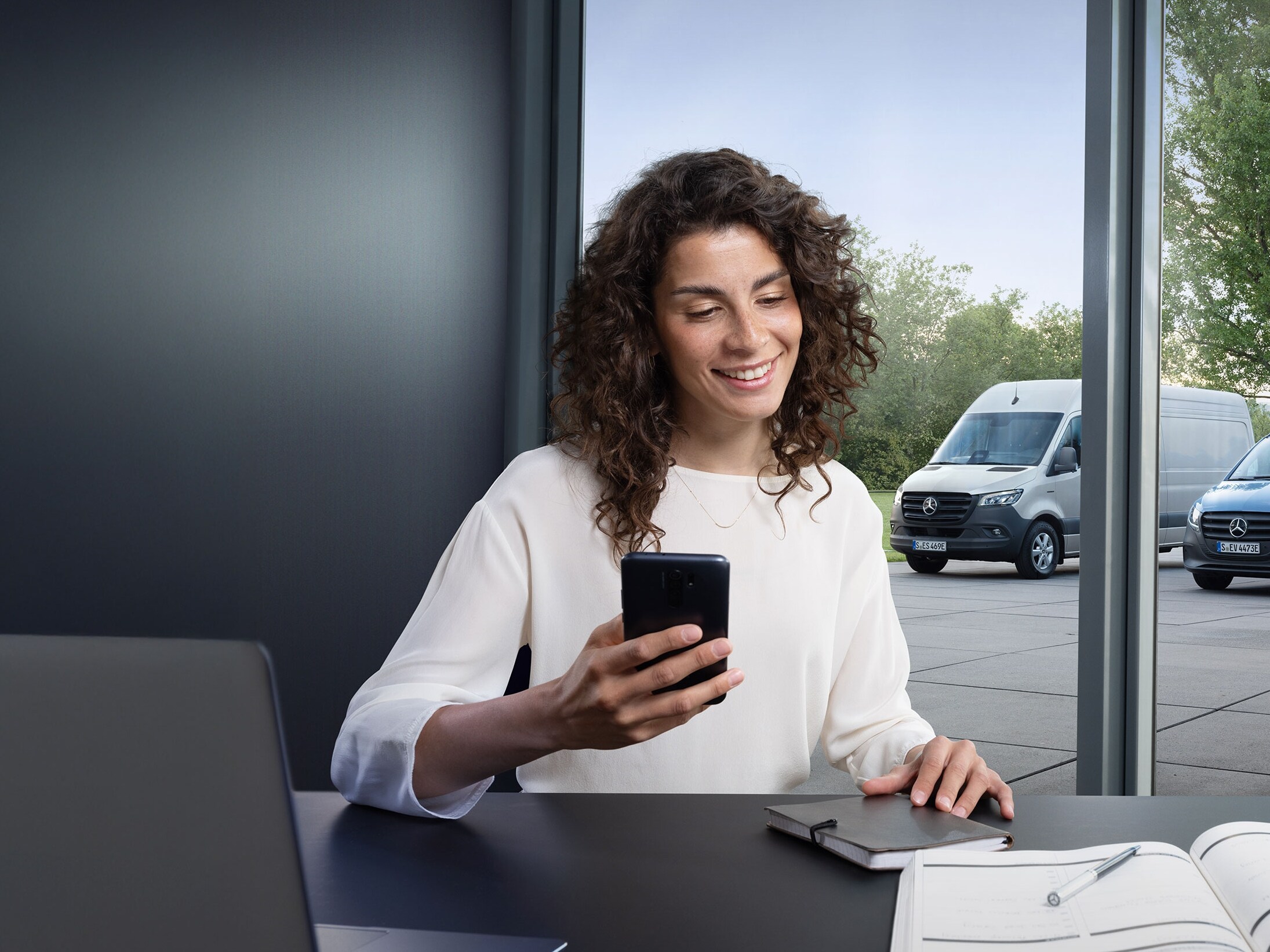 Mujer utilizando un smartphone para controlar el monitoreo de la flota de varios furgones Mercedes-Benz en la ventana de la oficina.