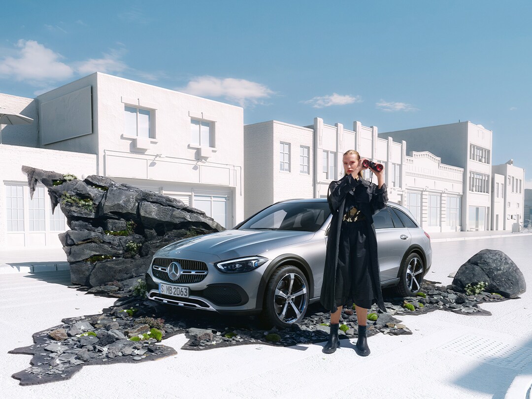 visual lateral de un Clase C Estate All-Terrain en color gris con una chica rubia vestida negra delante de él cogiendo unos prismáticos