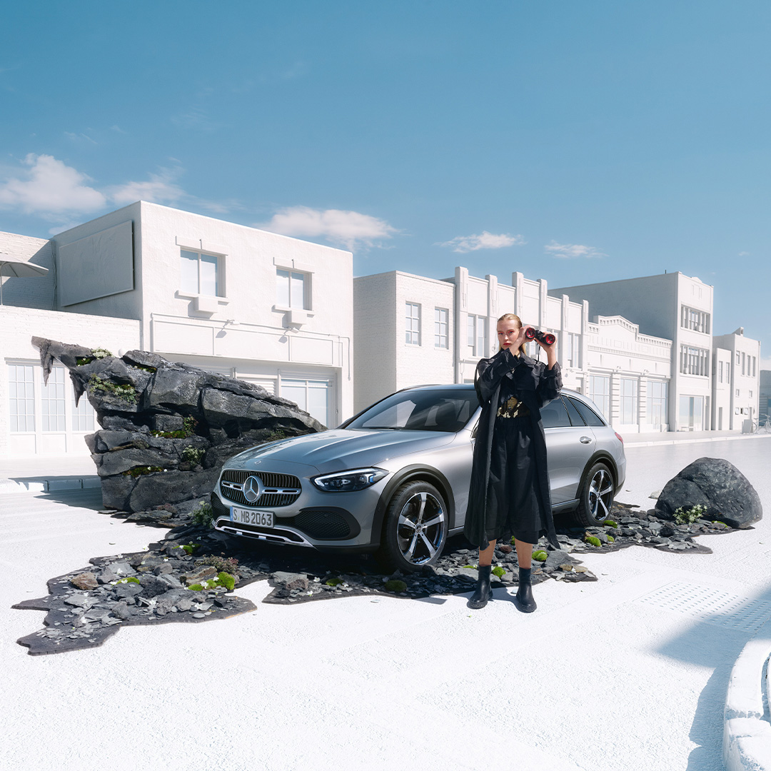 Oferta | Clase C Estate All-Terrain | Mercedes-Benz visual lateral de un Clase C Estate All-Terrain en color gris con una chica rubia vestida negra delante de él cogiendo unos prismáticos