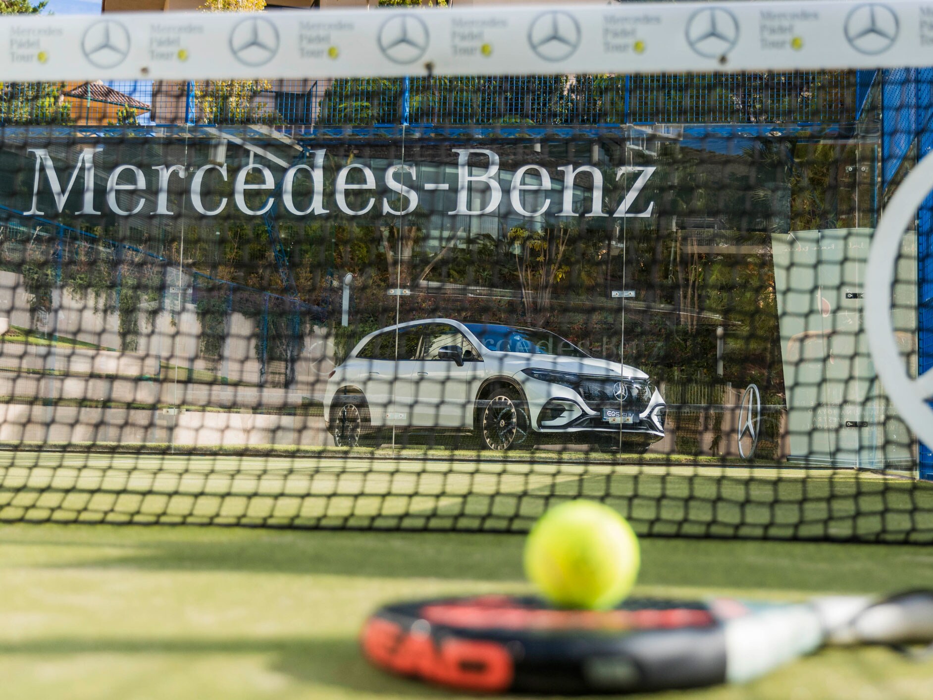 Visual raqueta y pelota sobre un campo de pádel y al fondo un EQS SUV de Mercedes-Benz