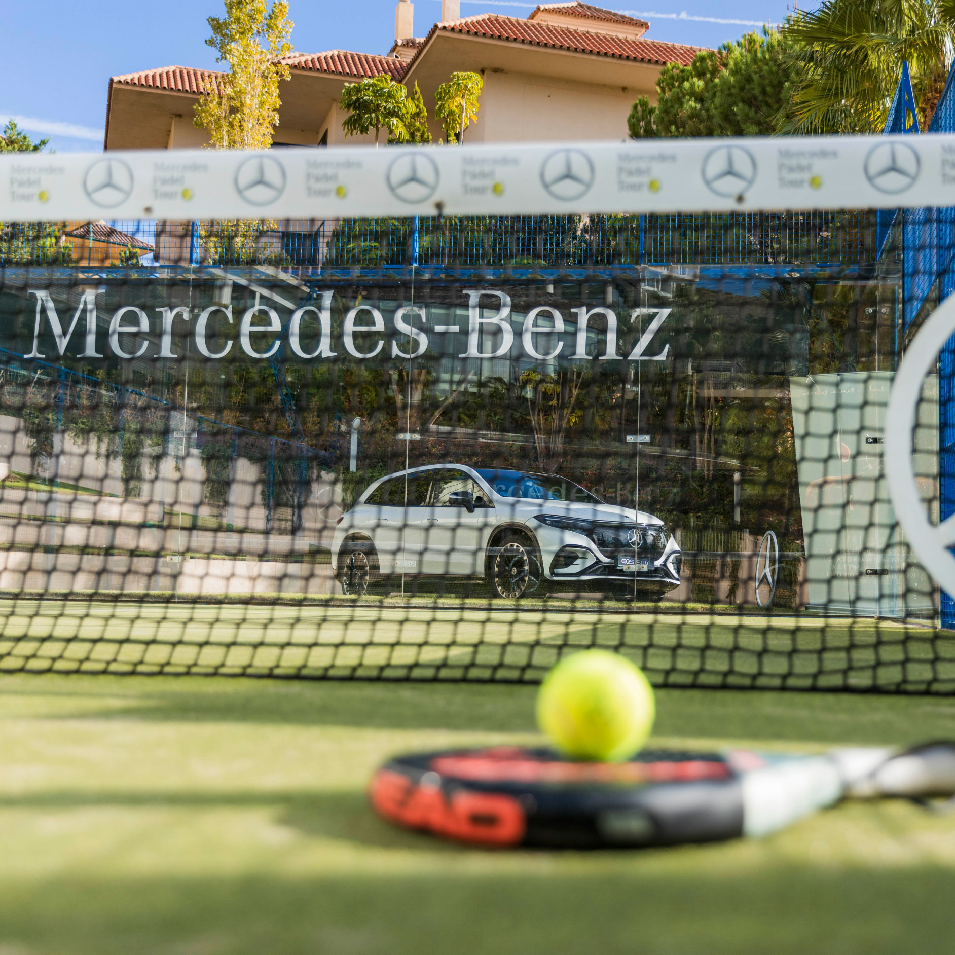 Pádel | Mercedes-Benz Visual raqueta y pelota sobre un campo de pádel y al fondo un EQS SUV de Mercedes-Benz