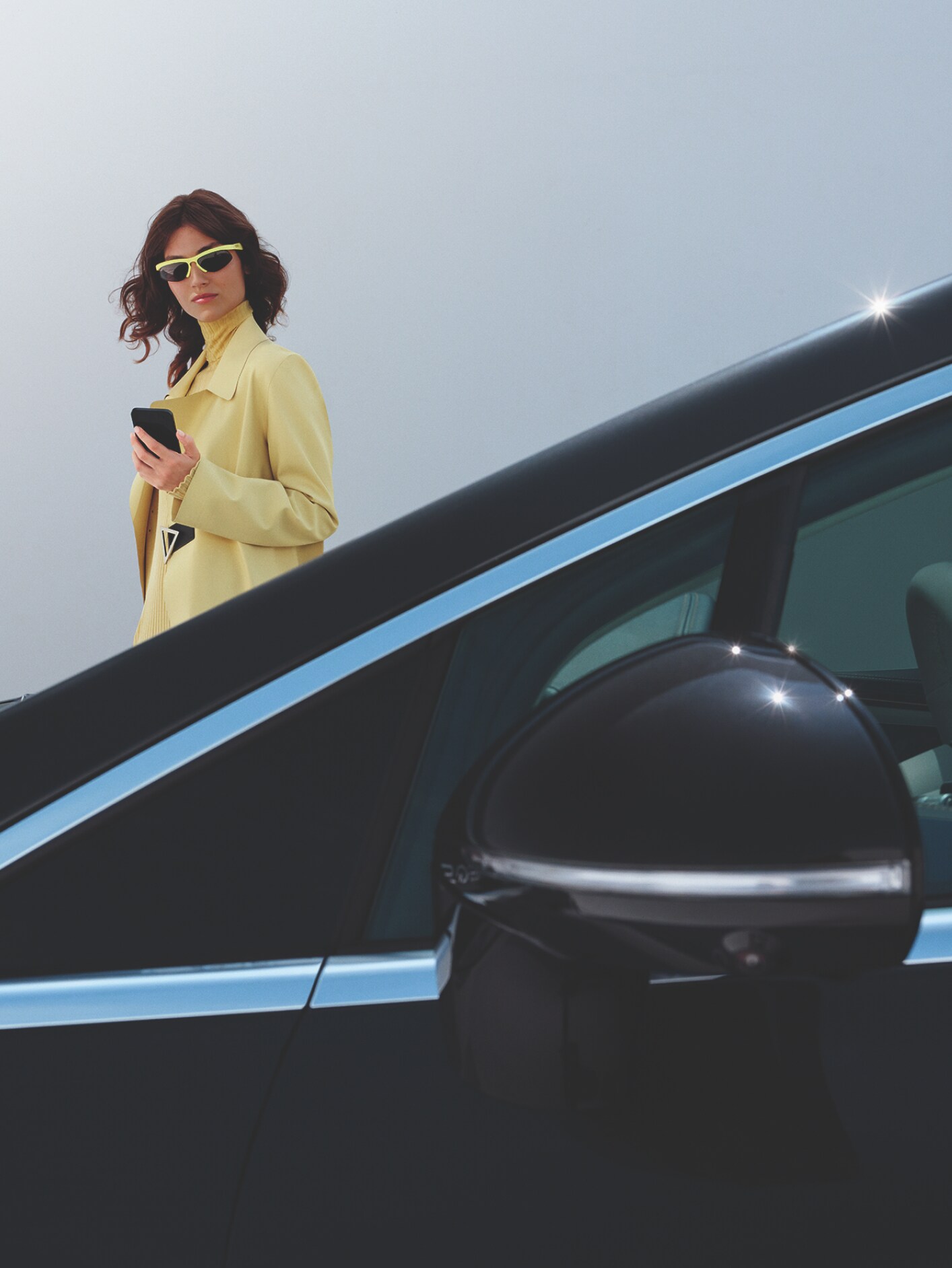 visual de una mujer vestida de amarillo con gafas de sol detrás de un coche negro