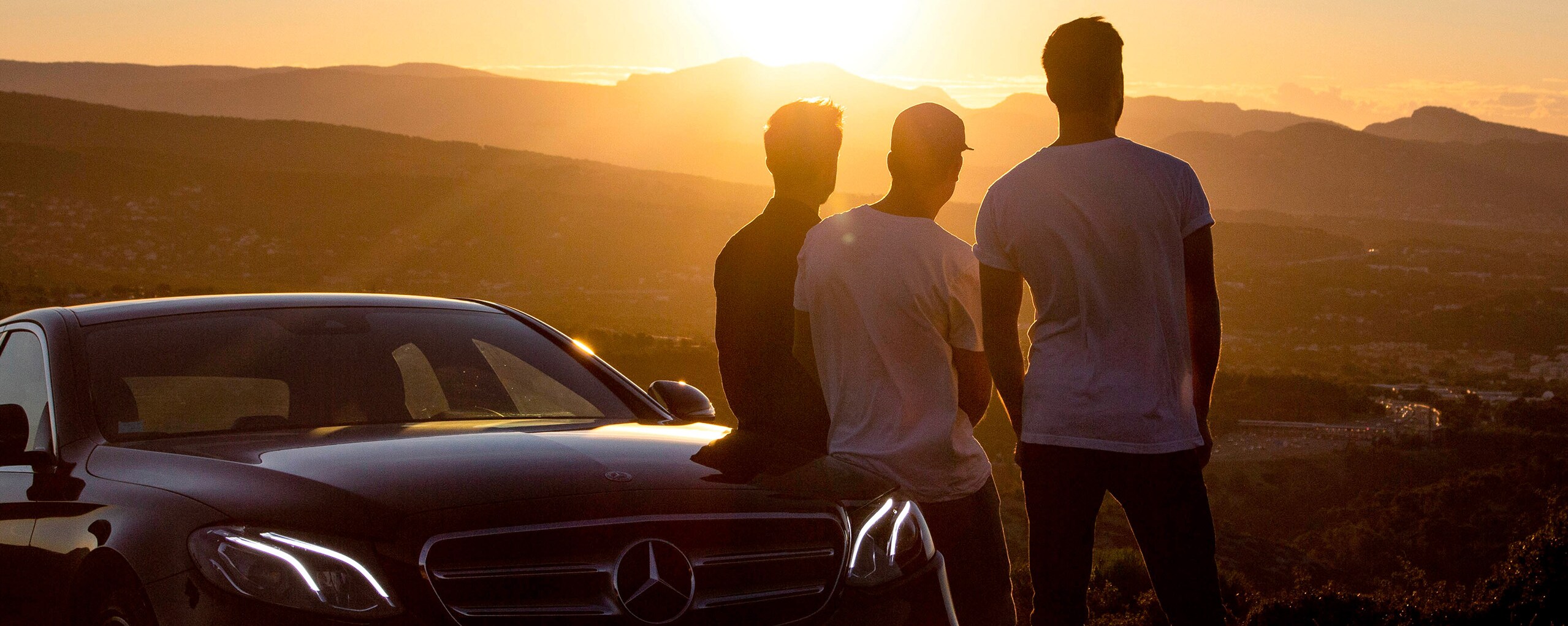Vista de tres hombres de espalda mirando el atardecer, apoyados en un Mercedes-Benz