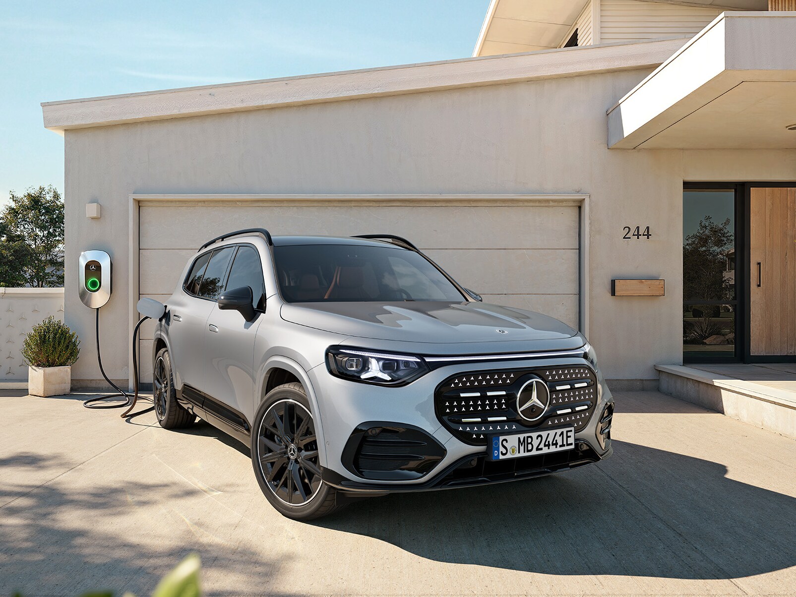 La imagen muestra un GLB eléctrico gris enchufado al cargador y aparacado fuera de una casa