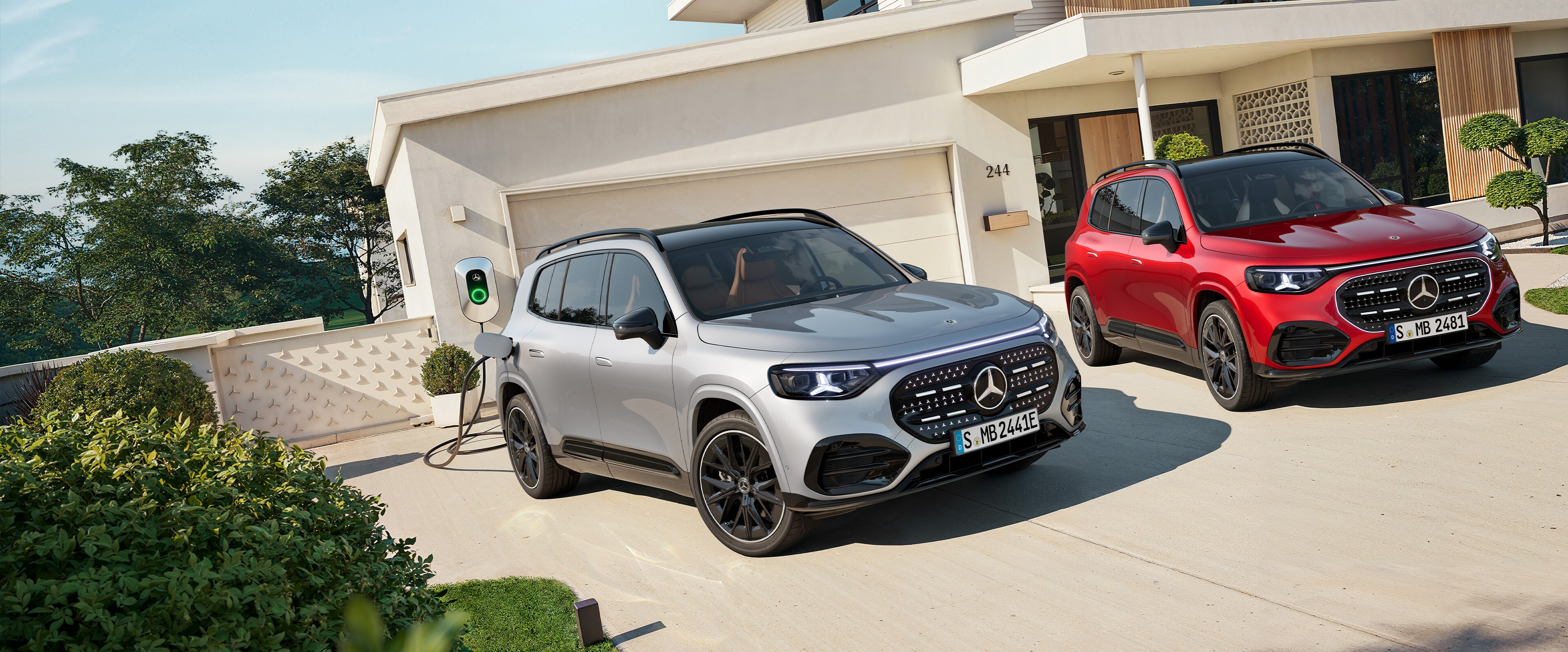 Un GLB eléctrico gris enchufado al cargador y otro rojo aparcados fuera de una casa