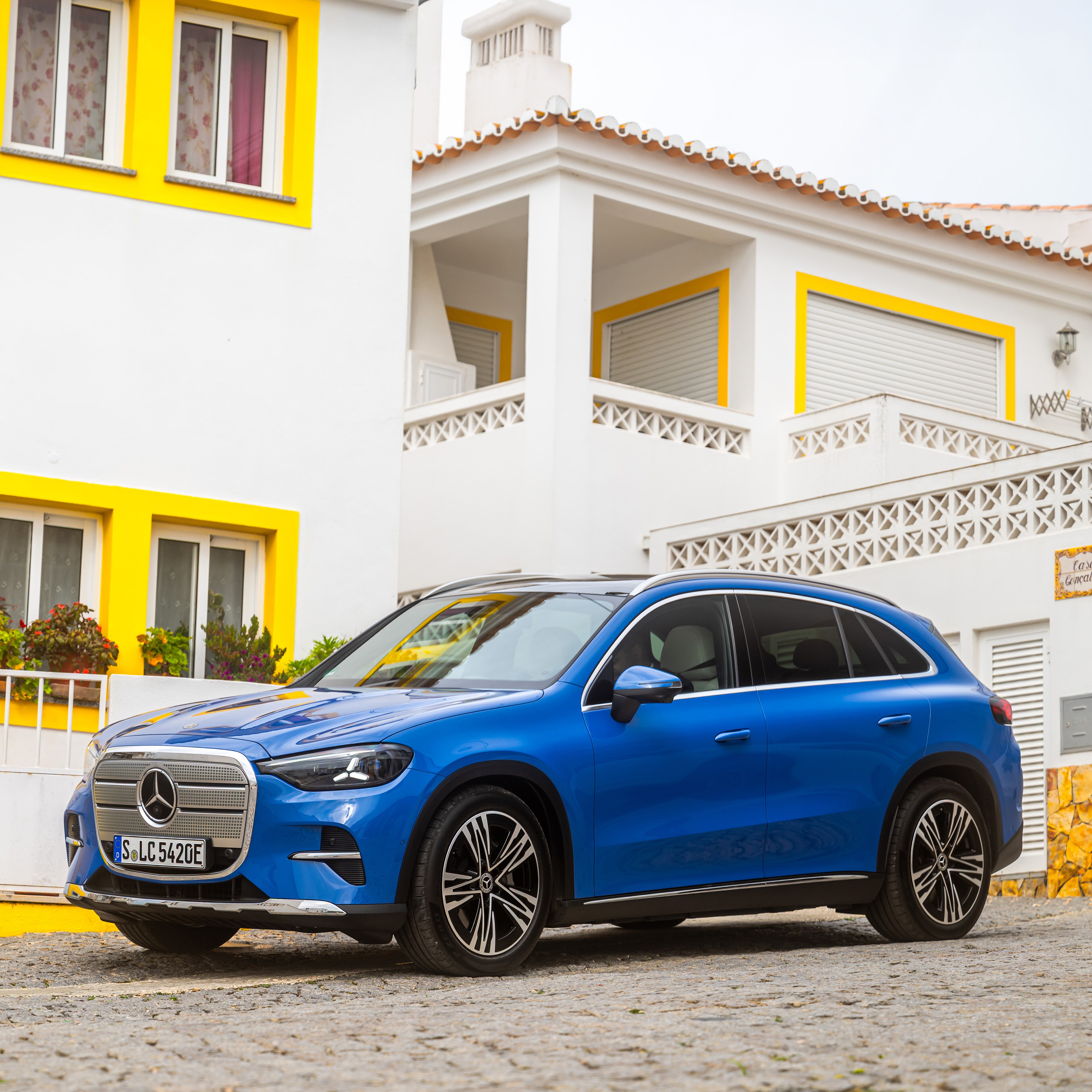 visual de un GLC en color azul en una plaza de suelo de piedra y enfrente de una casa con ventanas amarillas