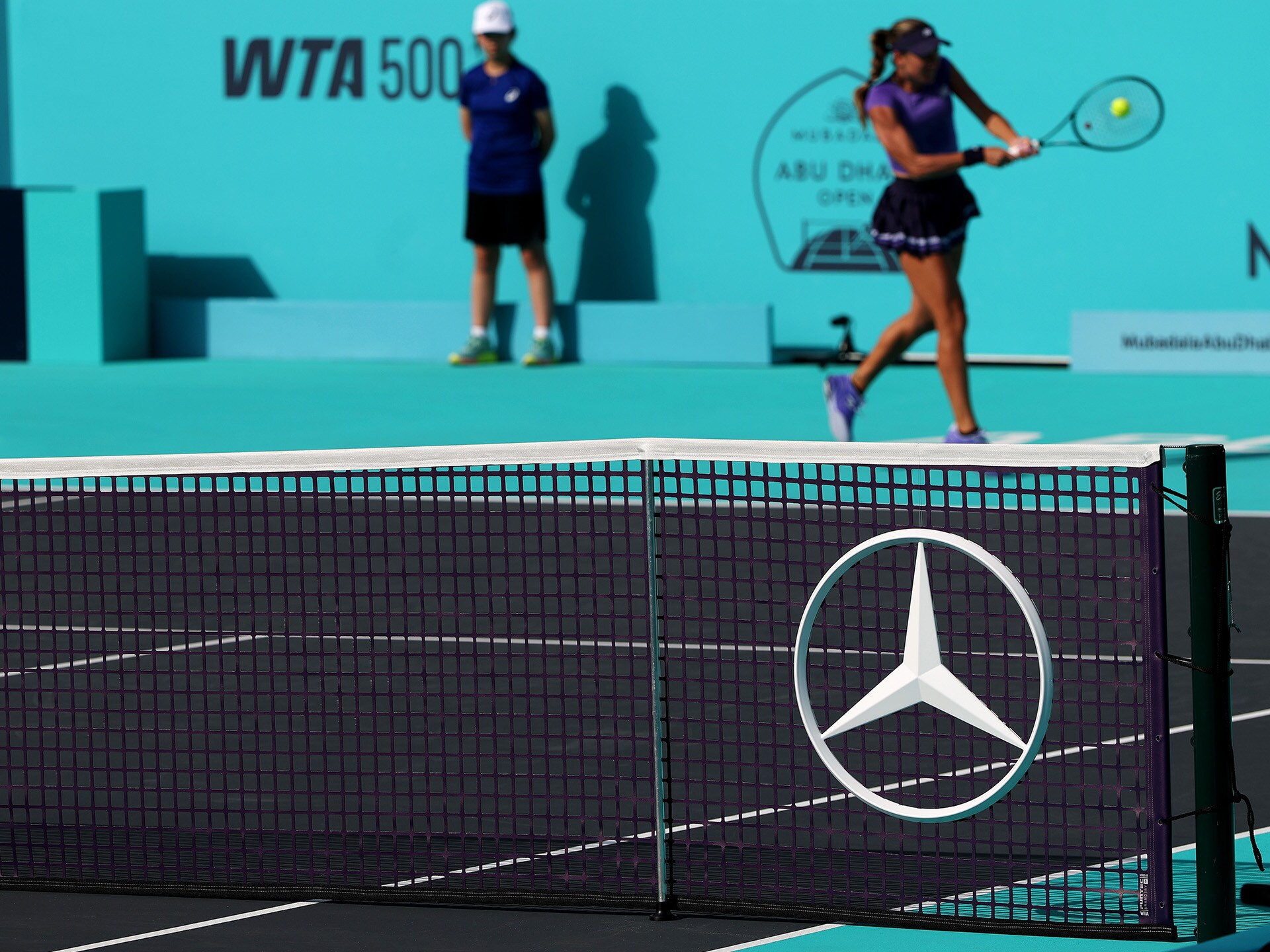 visual de una jugadora de tenis dándole a la pelota con una red de tenis en primer plano con el logo de Mercedes-Benz