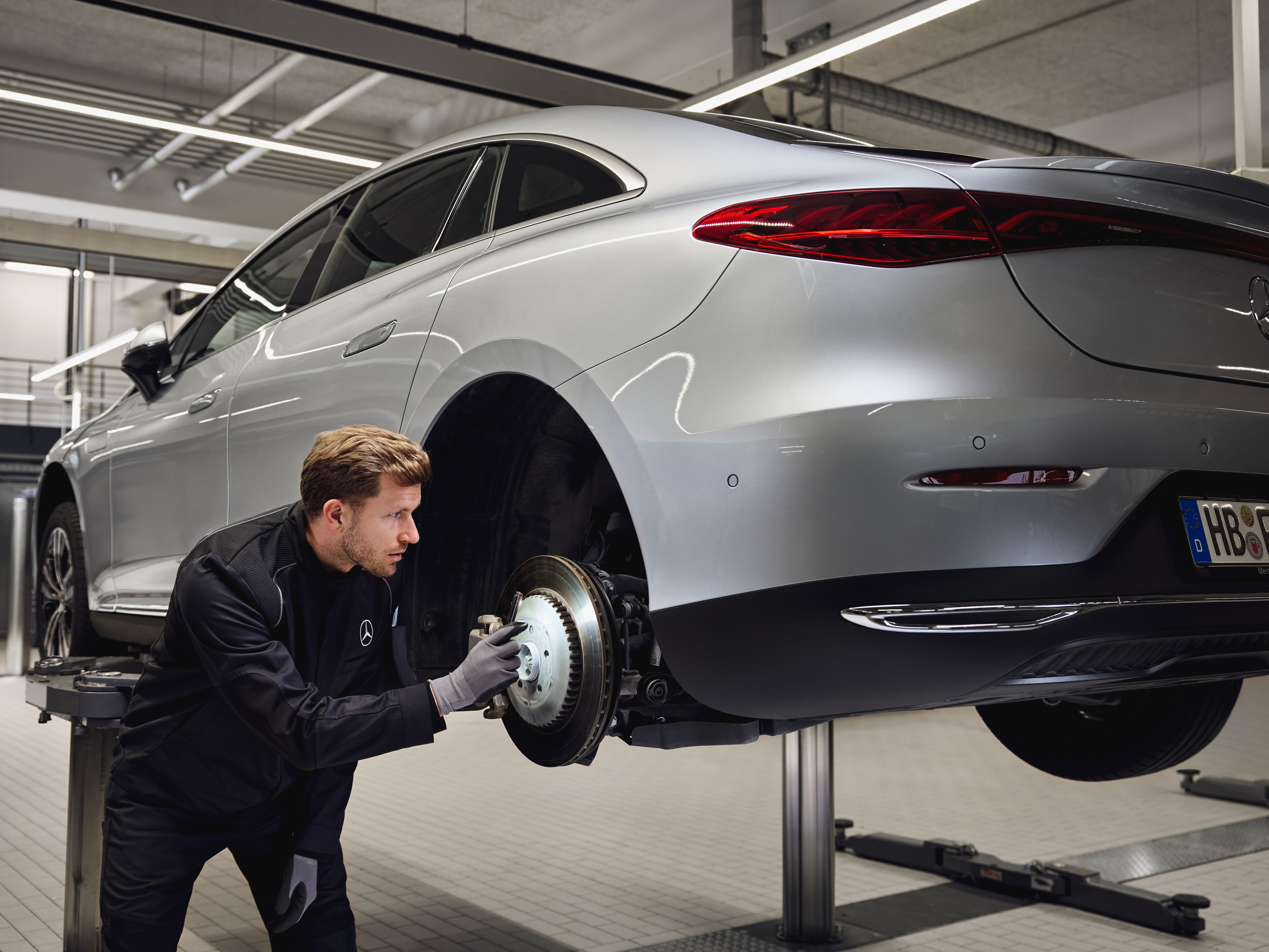 Un vehículo Mercedes-Benz elevado sobre una plataforma con un técnico inspeccionando el sistema de frenos traseros para un reemplazo con piezas originales.