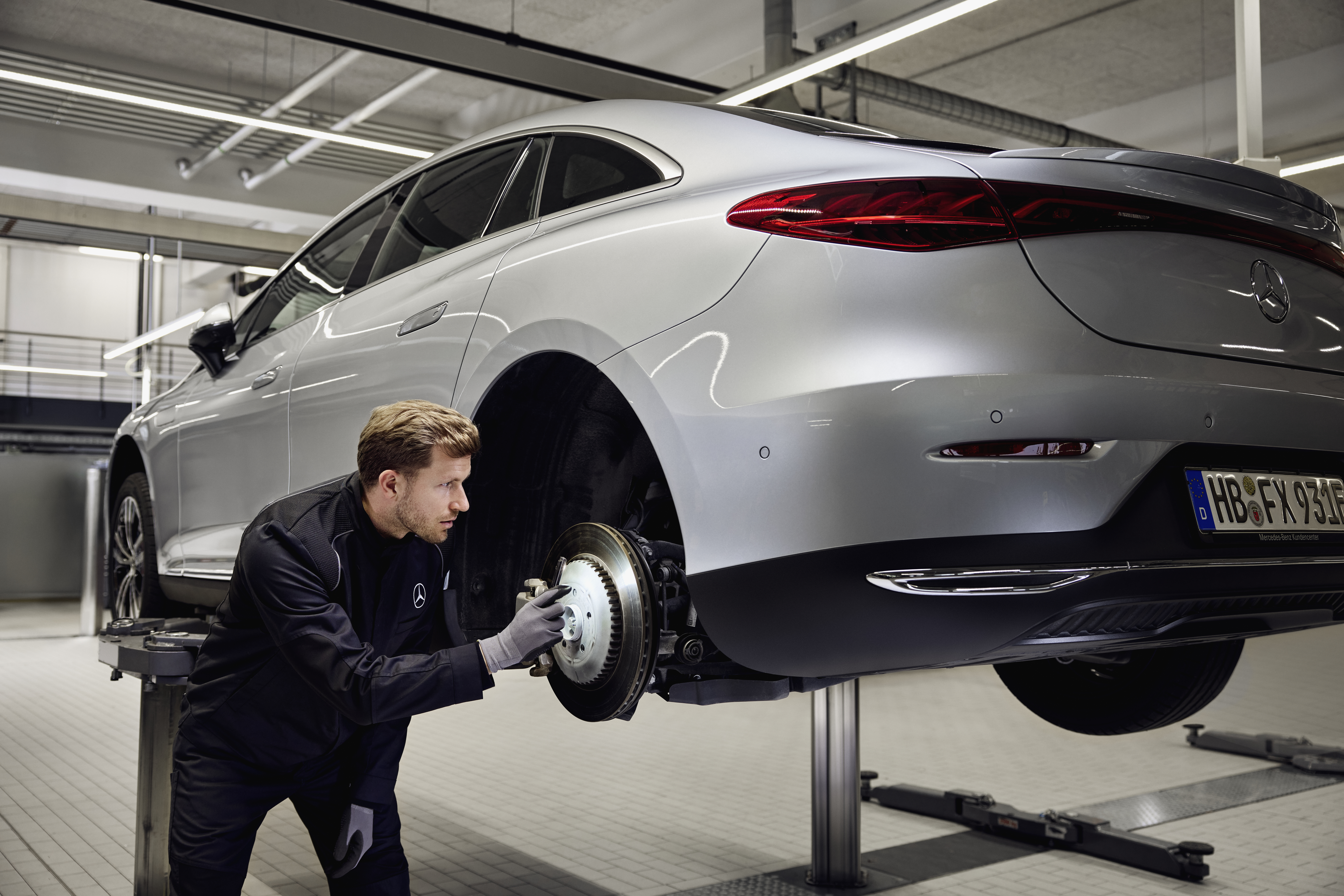 Recambio de frenos originales Mercedes-Benz Un vehículo Mercedes-Benz elevado sobre una plataforma con un técnico inspeccionando el sistema de frenos traseros para un reemplazo con piezas originales.