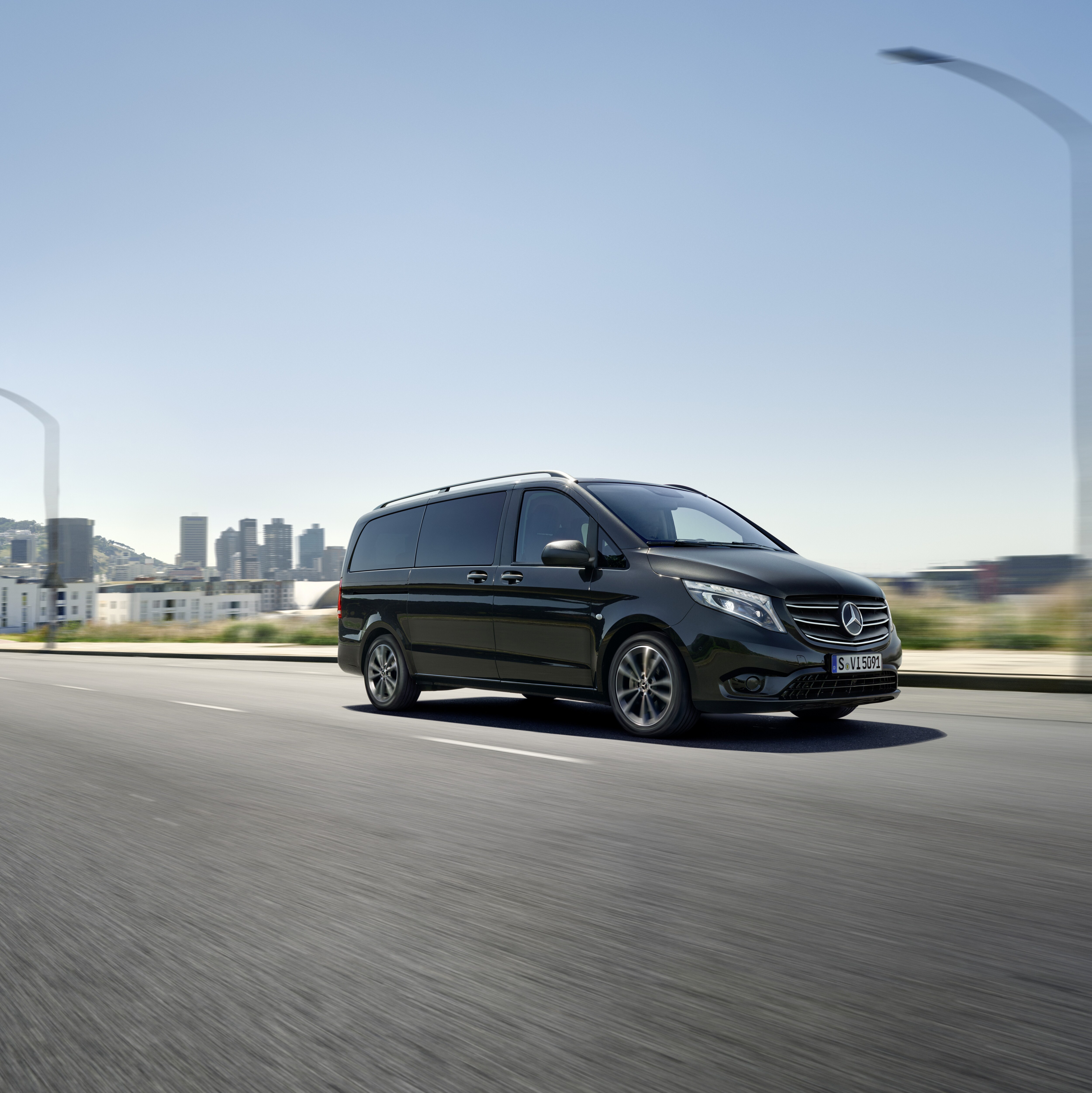 Mercedes-Benz Vito Tourer negra circulando por una carretera urbana, con edificios y montañas al fondo.